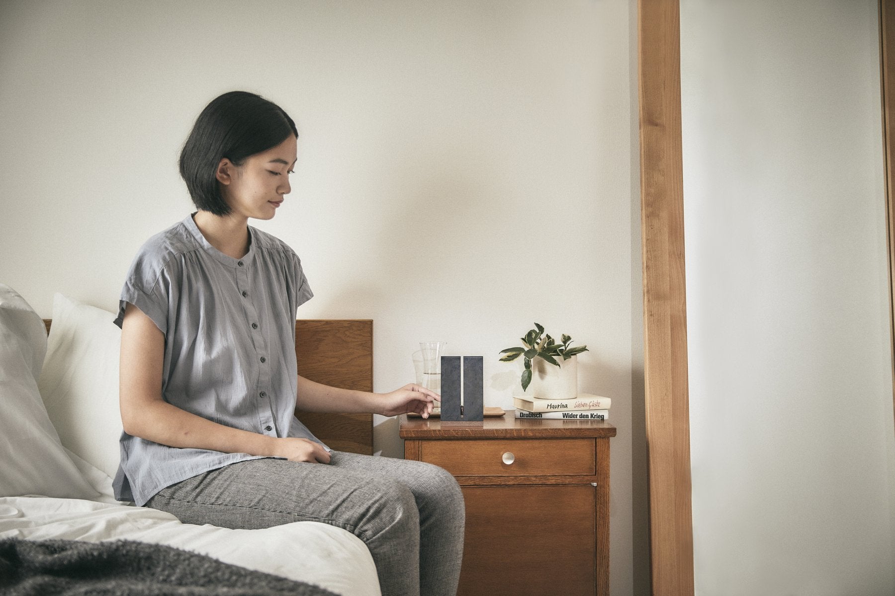 寝室でベッドに座る女性が、ナイトテーブルの上のHalocline Tallに手を伸ばしている生活シーン。観葉植物や本とともに