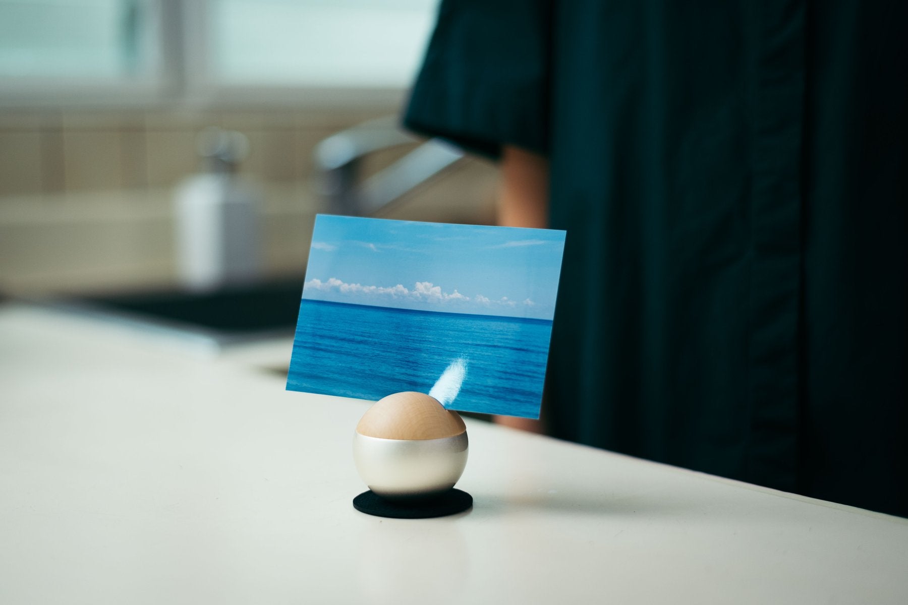 キッチンカウンターで、海の写真を立てた木目調のPictuary Sphereを置いた生活シーン。奥に人物が写る