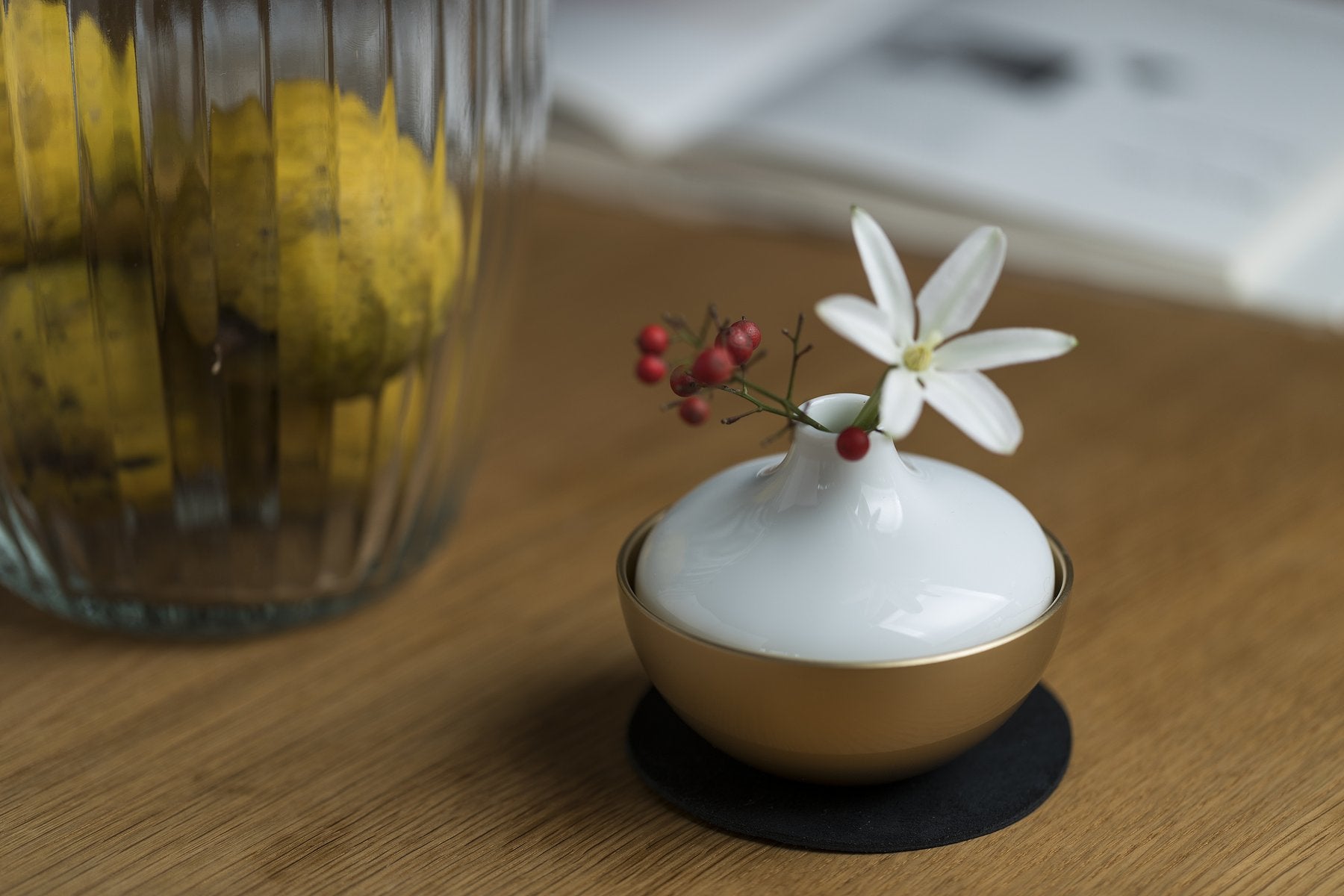 木製テーブルの上で、金色のChering(花立)に白い花器を重ねて一輪挿しにした様子。奥には瓶入りの柑橘と本が写る